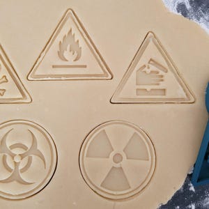 May include: A close-up of a baking sheet with five different cookie cutters. The cutters are shaped like warning signs, including a skull and crossbones, a biohazard symbol, a radiation symbol, a fire symbol, and a hand in a warning sign. The cookie cutters are made of blue plastic.