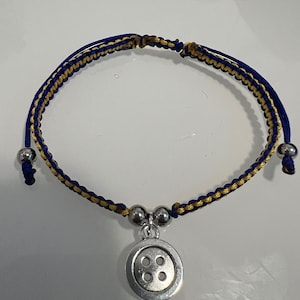 May include: A blue and gold braided bracelet with a silver button charm. The button has four holes.