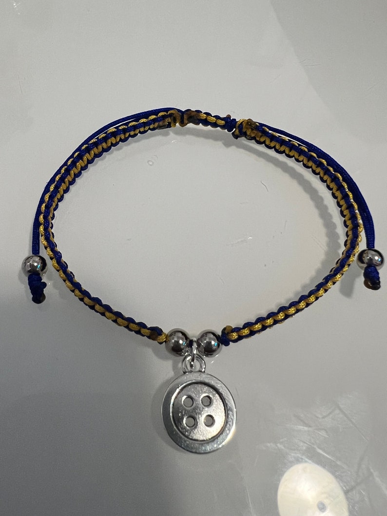 May include: A blue and gold braided bracelet with a silver button charm. The button has four holes.