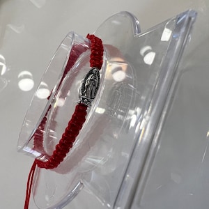 May include: Red string bracelet with a silver charm featuring a religious figure. The bracelet is displayed on a clear plastic stand.