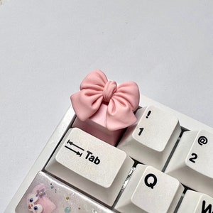 May include: A light pink bow-shaped keycap sits atop a white keyboard key. The keycap is a light pink color and is shaped like a bow. The keyboard keys are white with black lettering, including the "Tab" key.