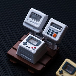 May include: A miniature white gaming console with a brown base. The console has a screen that reads "GAME" and a joystick. There are two other white devices attached to the console, one with a screen and one with a calculator-like keypad.