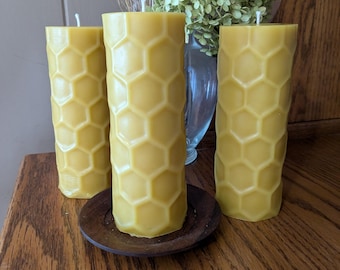 May include: Three yellow beeswax pillar candles with a honeycomb pattern, standing on a small wooden plate. The candles are cylindrical and have a natural, textured appearance. A vase of dried flowers is in the background.