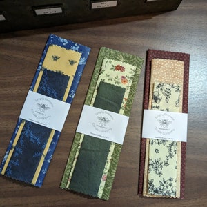 May include: Three sets of beeswax wraps, each with a different pattern, are displayed on a wooden surface. The wraps are bundled with a white label that reads "Simply Beeswax Candle Company." The patterns include bees, floral, and polka dots.