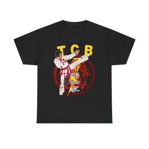 Puede incluir: Camiseta negra con un gráfico de una figura en posición de kárate, con las letras "TCB" en amarillo. La figura lleva un uniforme de kárate blanco con cinturón rojo. Un dragón rojo y caracteres chinos en el fondo.