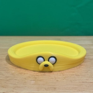 May include: Yellow plastic coaster with a cartoon dog face. The dog has black eyes, a black nose, and a black mouth.