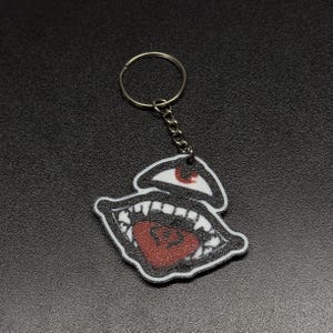 May include: A keychain with a graphic design of an open mouth with sharp teeth and a red heart-shaped tongue. Above the mouth is an eye with a red iris. The keychain has a silver ring and chain.