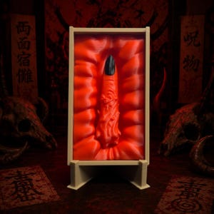 May include: A rectangular beige frame containing a sculpted orange finger with a black fingernail, set against a rippled orange background. The finger features intricate, textured details along its length, and the entire piece is displayed on a stand.