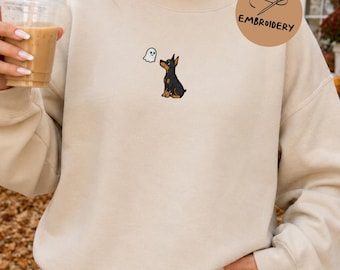 May include: A beige crewneck sweatshirt featuring a small embroidered graphic on the chest of a black and tan dog looking up at a tiny white ghost. The sweatshirt has long sleeves and a ribbed neckline, cuffs, and hem.