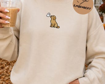 May include: A beige crewneck sweatshirt featuring a small embroidered design of a golden-coloured dog looking up at a tiny white ghost. The sweatshirt has long sleeves and a ribbed neckline, providing a simple and clean look for casual daily wear.