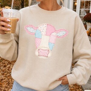 May include: A beige long-sleeve sweatshirt featuring a central graphic of a cow head composed of various pastel-coloured patchwork patterns, including pink and blue gingham and floral prints, with visible stitched outlines on the fabric.