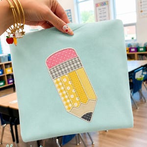 May include: A light mint green sweatshirt featuring a large, central applique of a pencil. The pencil design is constructed from various patterned fabric patches in shades of yellow, pink, grey, and black, with a stitched outline.