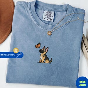 May include: A blue jean colored Comfort Colors t-shirt with an embroidered design of a German Shepherd puppy looking at a butterfly. The shirt has a round neckline and short sleeves. A gold necklace with a pendant is also visible.