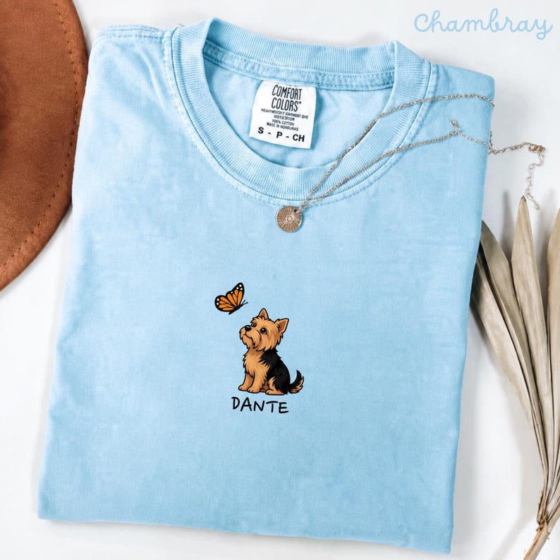Folded CHAMBRAY Comfort Colors shirt with cute Yorkshire Terrier puppy and butterfly graphic, personalized dog lover tee with custom pet name, minimalist pet shirt flat lay aesthetic gift idea.