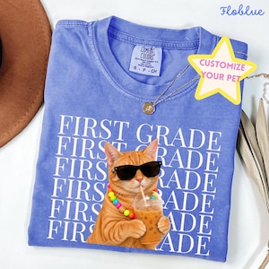 May include: A periwinkle blue crew neck t-shirt featuring a repeating white text pattern that reads FIRST GRADE. Centered is a graphic of an orange tabby cat wearing black sunglasses and a colorful beaded necklace while holding an iced coffee.