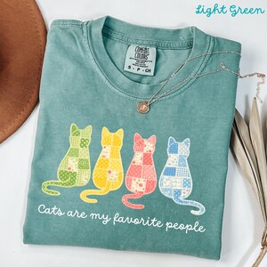 Peut inclure: Un t-shirt vert clair orné d'un motif de quatre chats avec un design en patchwork aux tons vert, jaune, rose et bleu. Sous les chats, le texte indique "Cats are my favorite people". Le t-shirt présente un col rond et une étiquette Comfort Colors.