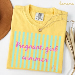 Banana yellow Comfort Colors shirt with vertical mint green stripes and pink script text reading “Pregnant girl summer” centered across the front, with a small pink sun icon detail at the bottom corner.