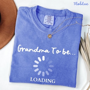 May include: A periwinkle blue short-sleeved t-shirt featuring the white text "Grandma To be..." above a circular loading icon and the word "LOADING" in white. The shirt has a Comfort Colors brand label at the inner neckline and a crew neck collar.