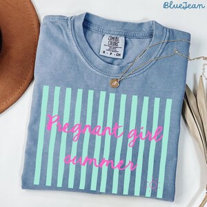 blue jean Comfort Colors shirt with vertical mint green stripes and pink script text reading “Pregnant girl summer” centered across the front, with a small pink sun icon detail at the bottom corner.