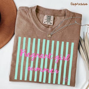 Espresso Comfort Colors shirt with vertical mint green stripes and pink script text reading “Pregnant girl summer” centered across the front, with a small pink sun icon detail at the bottom corner.
