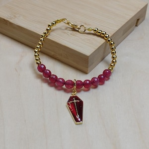 May include: Gold beaded bracelet with a strand of red gemstone beads. A gold charm of a red coffin with a gold cross hangs from the bracelet. The bracelet is displayed on a light wood surface.