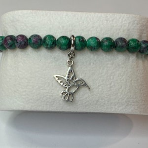 May include: A green and pink gemstone bracelet with a silver hummingbird charm.