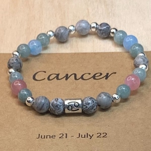 May include: A Cancer zodiac bracelet with grey, blue, and pink beads. The bracelet has a silver charm with the Cancer zodiac symbol. The bracelet is on a brown background with the text "Cancer" and "June 21 - July 22".