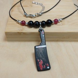 May include: A black necklace with red and black beads and a pendant shaped like a bloody butcher knife.