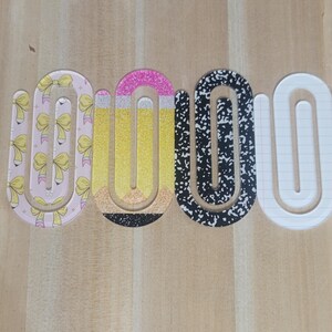 May include: A set of decorative paper clips in a U-shape. The clips feature various designs, including bows, a pencil, a black and white speckled pattern, and a lined paper design. The clips are arranged on a light brown wooden surface.