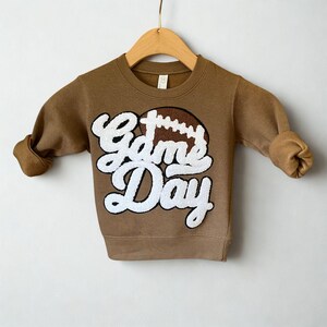 以下が含まれることがあります： 「Game Day」の文字とフットボールのグラフィックが白で描かれた茶色のスウェットシャツ。袖は折り返されています。スウェットシャツは木製のハンガーに吊るされています。