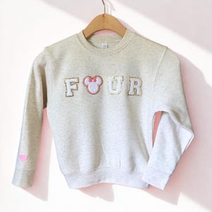 K&ouml;nnte beinhalten: Hellgraues Sweatshirt mit dem Wort "FOUR" in goldglitzernden Buchstaben und einer rosa Minnie-Mouse-Schleife. Auf dem linken &Auml;rmel ist ein rosa Herz gestickt. Das Sweatshirt h&auml;ngt an einem Holzb&uuml;gel.