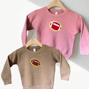 以下が含まれることがあります： 子供用のスウェットシャツ2枚。1枚はダスティーローズ、もう1枚は茶色で、それぞれにフットボールのアップリケが付いています。フットボールは赤色で、白いステッチと黄色の縁取りが施されています。どちらも長袖で、木製のハンガーに吊るされています。