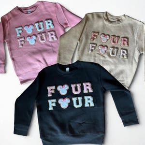 K&ouml;nnte beinhalten: Drei Sweatshirts mit Rundhalsausschnitt in Rosa, Beige und Schwarz. Jedes Sweatshirt zeigt das Wort "FOUR" in einem Blockbuchstabenstil mit einem Mickey Mouse-Kopf in der Mitte. Die Buchstaben und M&auml;usek&ouml;pfe sind in einer Kontrastfarbe gehalten.