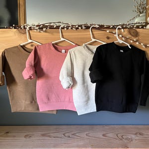 K&ouml;nnte beinhalten: Vier Sweatshirts mit Rundhalsausschnitt in den Farben Braun, Rosa, Hellgrau und Schwarz. Jedes Sweatshirt h&auml;ngt an einem wei&szlig;en Kleiderb&uuml;gel, die &Auml;rmel sind hochgekrempelt. Die Sweatshirts werden auf einem Holzregal und einer dunkelblauen Wand pr&auml;sentiert.