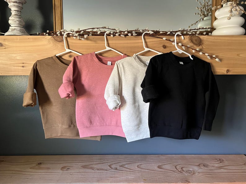 K&ouml;nnte beinhalten: Vier Sweatshirts mit Rundhalsausschnitt in den Farben Braun, Rosa, Hellgrau und Schwarz. Jedes Sweatshirt h&auml;ngt an einem wei&szlig;en Kleiderb&uuml;gel, die &Auml;rmel sind hochgekrempelt. Die Sweatshirts werden auf einem Holzregal und einer dunkelblauen Wand pr&auml;sentiert.