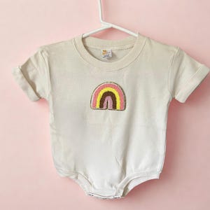 Puede incluir: Un body de bebé color crema con mangas cortas, colgado de una percha blanca. El body presenta un diseño de arcoíris bordado colorido en tonos de rosa, amarillo y marrón. El fondo es rosa liso.