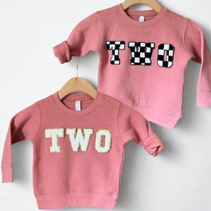 Könnte beinhalten: Zwei rosa Sweatshirts für Kinder. Eines zeigt das Wort „TWO“ in goldumrandeten weißen Buchstaben, das andere das gleiche Wort in schwarz-weiß karierten Buchstaben. Beide hängen an Holzbügeln.