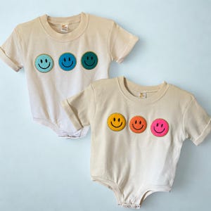 Peut inclure: Deux bodies de bébé beiges avec des manches retroussées. Chaque body présente trois visages souriants brodés en rangée. L'un a des visages bleus, turquoise et verts, l'autre des visages jaunes, oranges et roses. Les bodies ont un col rond et des fermetures à boutons-pression.