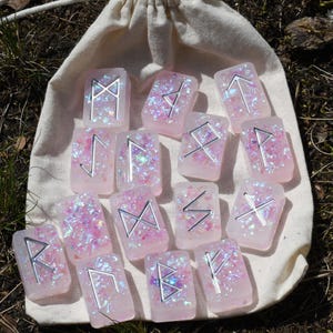 Peut inclure: Un ensemble de 15 pierres de runes roses et irisées avec des runes argentées sur chaque pierre. Les pierres sont dans un sac en tissu blanc avec cordon.