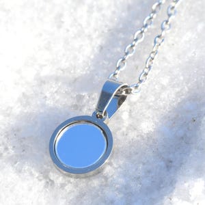 Peut inclure: Un collier argenté avec un pendentif circulaire. Le pendentif a un centre bleu clair et une surface polie et réfléchissante. La chaîne est délicate et le pendentif est suspendu à une petite bélière.