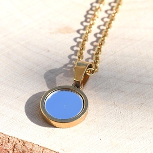 May include: A gold-colored necklace with a round pendant featuring a blue mirrored surface. The pendant is set in a gold-toned bezel and hangs from a delicate gold chain. The necklace is displayed on a light-colored wooden surface.