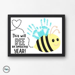 Puede incluir: Una impresión enmarcada con una abeja hecha con huellas de manos. La abeja es amarilla con rayas negras y tiene una cara sonriente. El texto "This will BEE an amazing YEAR!" está impreso debajo de la abeja.