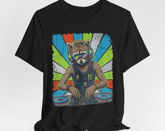 Puede incluir: Camiseta negra con un gráfico de mapache DJ. El mapache lleva gafas de sol, auriculares y una camiseta sin mangas con el número 13. El fondo es una explosión de colores azul, verde, naranja y blanco.