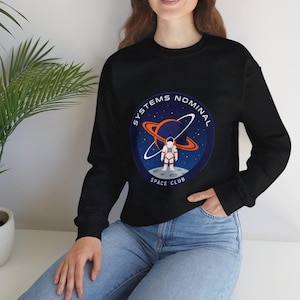 May include: Black crew neck sweatshirt with a circular graphic featuring an astronaut on a moon, a planet with rings, and the words "SYSTEMS NOMINAL SPACE CLUB". The design is in shades of blue, orange, and white.