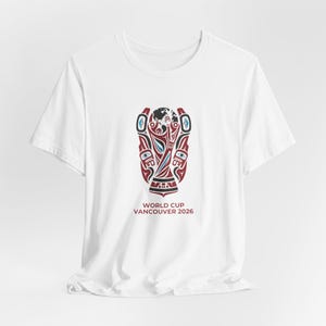 May include: White t-shirt featuring a stylized graphic in red, black, and blue. The graphic depicts a trophy with a globe and tribal art elements. The text "WORLD CUP VANCOUVER 2026" is printed below the graphic.