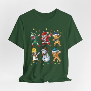 Op de afbeelding: Een donkergroen t-shirt met een kerstontwerp. Het toont personages als de kerstman, een elf, een rendier, een pinguïn, een sneeuwman en een peperkoeken man, allemaal aan het dabben. Kleurrijke lichtslingers zijn ook inbegrepen.