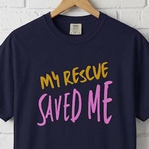 May include: A navy blue short-sleeve t-shirt featuring the text "MY RESCUE" in gold and "SAVED ME" in pink. The shirt has a crew neckline and is displayed on a wooden hanger, showcasing a casual design with bold, hand-lettered style typography.