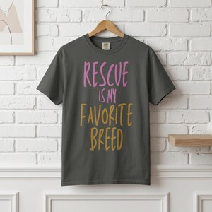 May include: A dark gray short-sleeve t-shirt featuring the text "RESCUE IS MY FAVORITE BREED" printed on the front. The word "RESCUE" is in pink, while the words "IS MY FAVORITE BREED" are in a mustard yellow color, all in a handwritten font.