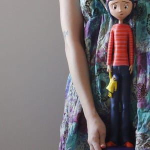 Puede incluir: Una figura de muñeca alta de una niña con cabello azul, que lleva una camisa de manga larga a rayas rojas y naranjas y pantalones azul oscuro. La muñeca sostiene una pequeña figura amarilla. La muñeca está sobre una base azul.