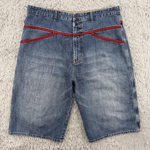 May include: Blue denim shorts with red detailing across the waist. The shorts feature a classic five-pocket design and a button and zip fastening. The denim has a faded wash, giving a vintage appearance.